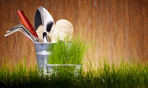 A close-up view of a garden scene featuring a small patch of vibrant green grass in the foreground, with a backdrop of a wooden fence. In the middle ground, there are two metallic gardening buckets; one contains various hand tools such as a trowel, pruning shears, and a hand rake, along with a ball of twine, while the other bucket holds a clump of fresh, bright green grass or herb sprigs. The outdoor environment is well-lit under natural daylight, suggesting a clear and mild weather condition. The scene emphasizes gardening activities, with a focus on garden maintenance supplies and lush lawn elements, typical of services provided by companies like Garden Maintenance Harrow in the Harrow area, to support ongoing outdoor care and landscaping projects.